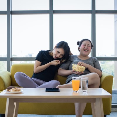 Overweight woman and asian girl enjoy eating food on sofa at home