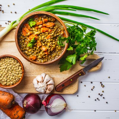 Lentil soup in a wooden bowl and ingredients on a white wooden background, top view.