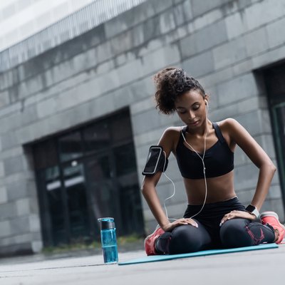 Young fitness woman in earphones sitting relaxed on yoga mat after workout with a water bottle outside