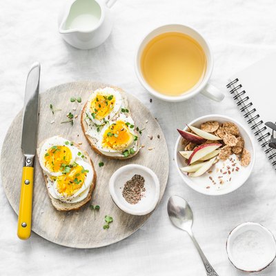 Morning breakfast table inspiration - sandwiches with cream cheese and boiled egg, yogurt with apple and flax seeds, herbal detox tea, notebook, glasses on light background, top view. Flat lay