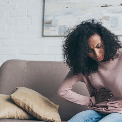 unhappy african american woman holding glass of water while sitting on sofa and suffering from stomach pain