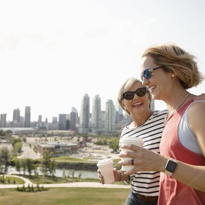 Smiling mother and daughter walking in sunny urban park with coffee