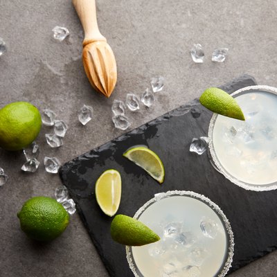 Top view of margarita cocktails with pieces of lime, ice cubes and wooden squeezer on grey tabletop