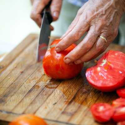 Close-up of Senior Adult Woman Slicing Tomato on Wooden Cutting Board - Stock Photo