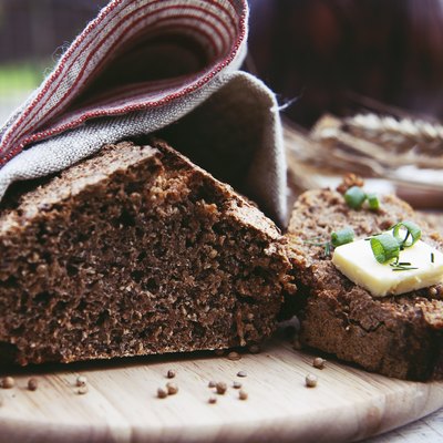 Loaf of freshly baked homemade whole grain rye bread with seeds cut in two on a wooden cutting board and covered with linen tea towel.