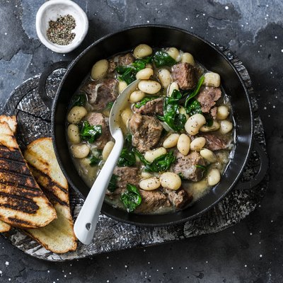 Beef, beans, spinach slow cooker stew in a pan on a wooden board on a dark background. Delicious homemade comfort food