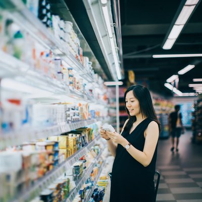Asian pregnant woman grocery shopping in supermarket and reading nutrition label on a packet of cheese