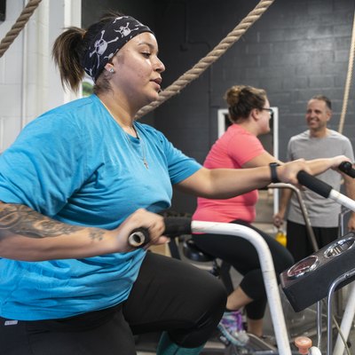 Two attractive body-positive women, Latino and Caucasian, doing a workout on the exercise bike in the gym under the supervision of the coach, the senior 55-years-old Cuban Hispanic man