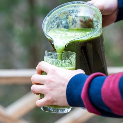 Closeup of behind, back of young man standing outside, outdoors, holding plastic blender container, pouring green vegetable, kale smoothie into drinking glass