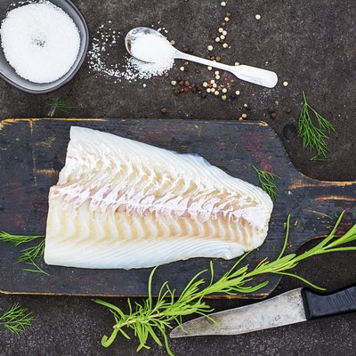 Raw cod before cooking on a black chopping Board with herbs and sea salt on a dark background. Top view