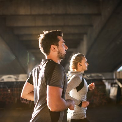 Young sporty man and woman with earphones running under the bridge outside in a city.