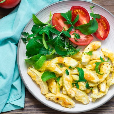 Slices of zucchini baked in spices with cheese on a plate with tomatoes and arugula on a wooden table