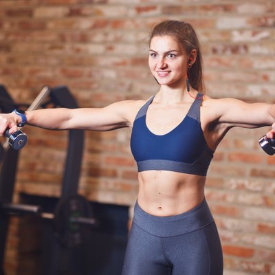 Portrait of attractive cheerful sportswoman with headphones in her ears. Sporty woman doing exercises with dumbbells for shoulders in the gym on the background of brick wall