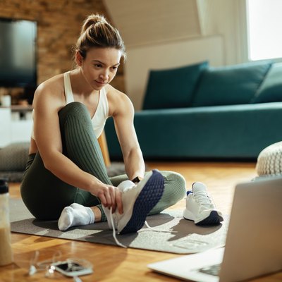 Athletic woman putting on sneakers while using laptop on the floor.