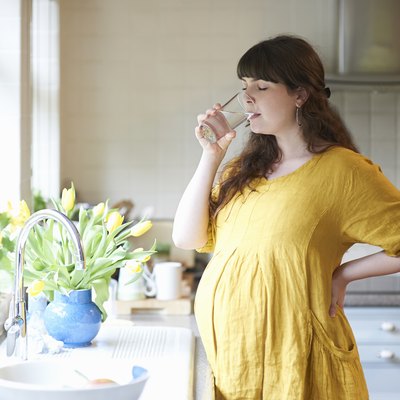 Pregnant woman drinking glass of water in kitchen at home.
