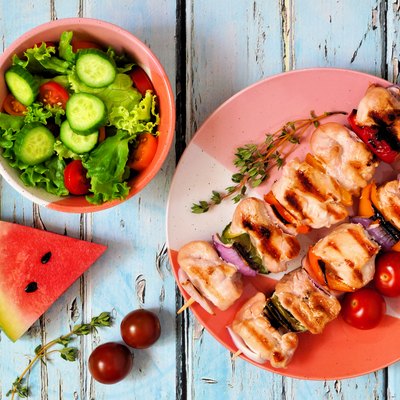 Grilled chicken and vegetable skewers with salad and watermelon, above scene over blue wood
