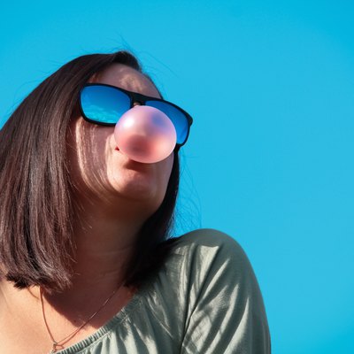 Low Angle View Of Woman Wearing Sunglasses Chewing Gum Blowing a Bubble Against Blue Background