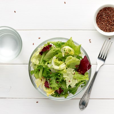Mixed salad in glass bowl with flax seed and water on white wooden table.