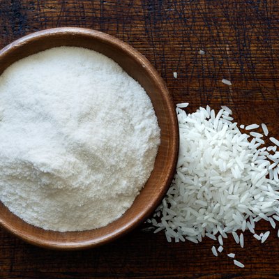 White rice flour in dark wooden bowl isolated on dark brown wood from above. Spilled rice.
