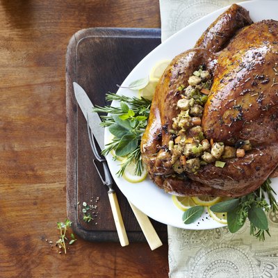 Holiday whole cooked turkey with glaze, herbs, and stuffing on fresh herbs and lemons on serving platter with carving utensils