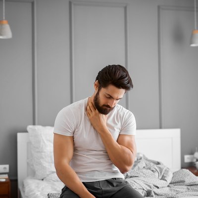 Portrait of annoyed handsome muscular bearded man having throat pain and sitting on the edge of the bed at home.