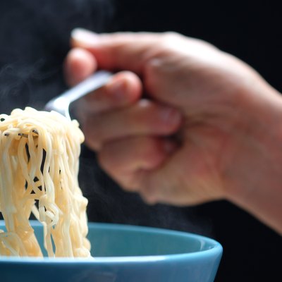 Hand holding fork full of hot noodles from soup in a blue bowl.
