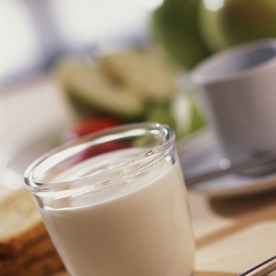 Glass of milk on table with cup, saucer, eating utensil and four slices of bread nearby, close-up, tilt, selective focus