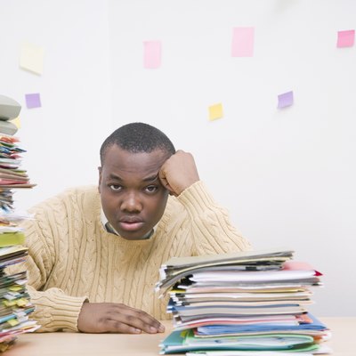 Man sitting next to pile of folders on desk, portrait, adhesive notes on wall in background