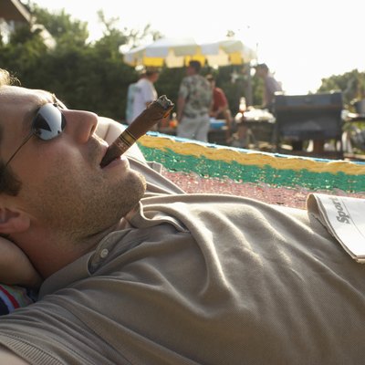 Man lying in hammock smoking cigar, friends by barbecue in background