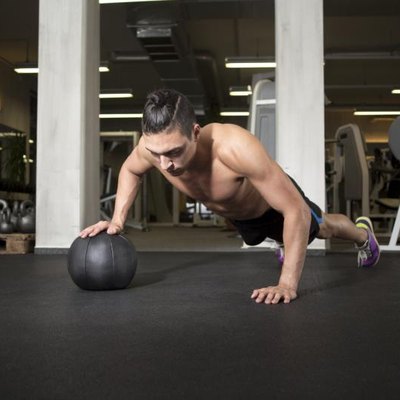 Fit and attractive athlete is performing a one-handed push-up on a medicine ball at the gym.