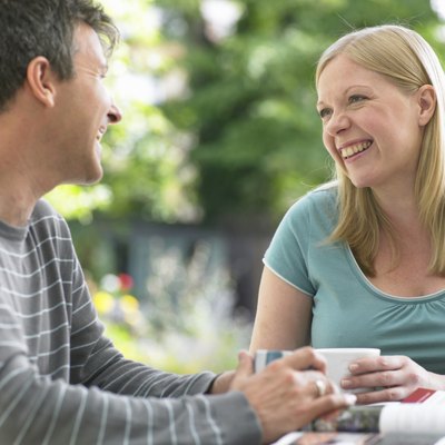 Couple sitting at table holding mugs, looking at each other, laughing