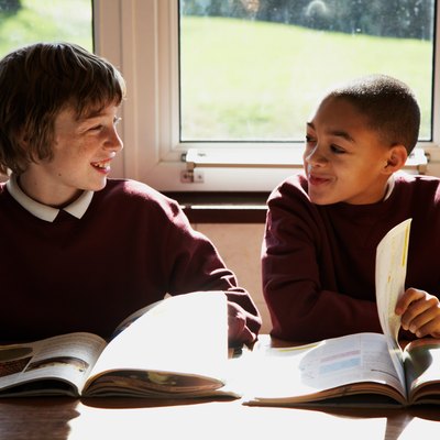 Two schoolboys (9-12) sitting in class turning pages in books, smiling