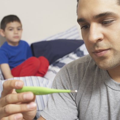 Close-up of a young man holding a digital thermometer with his son sitting on the bed behind him