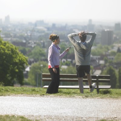 Man and woman resting after exercises, man stretching looking at cityscape, woman holding mobile phone