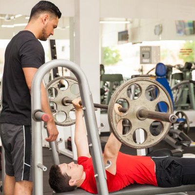 Profile view of a personal trainer spotting a young man while he lifts a barbell on the bench press at a gym