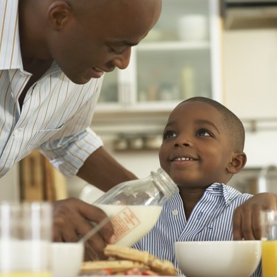 Father pouring milk in bowl for son (5-7) at breakfast table, smiling