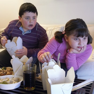 Overweight Brother and Sister Sitting Side by Side on a Sofa Eating Takeaway Food and Watching the TV