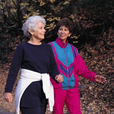 an elderly woman wearing black workout clothes walks with a middle-aged woman wearing red and blue workout clothes on a path covered with dead and fallen leaves