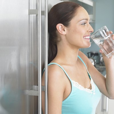 Young woman leaning against fridge, drinking glass of water, side view