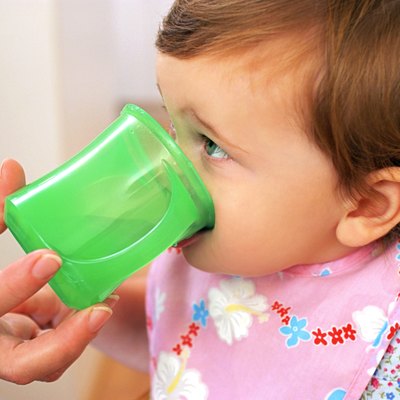 A baby girl drinks from a green cup with help from her mother.