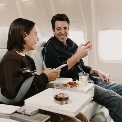 Man Sits in a Plane Seat Next to His Son, Sharing Headphones and Eating a Snack