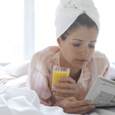 Young woman lying on bed reading newspaper, towel around head