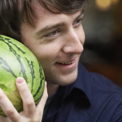 Young man holding watermelon up to ear, smiling, close-up