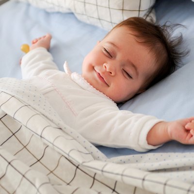 Baby smiling in bed with eyes closed and arms out.