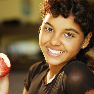 Close-up of a teen girl smiling at the viewer while holding the apple that she is eating.