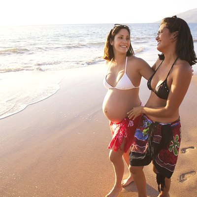 Two female friends walk in swimsuits along the beach; one woman is pregnant.
