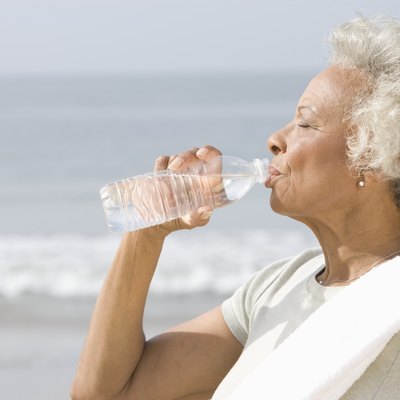 Senior woman stands with drinks water with a towel over her shoulder on beach