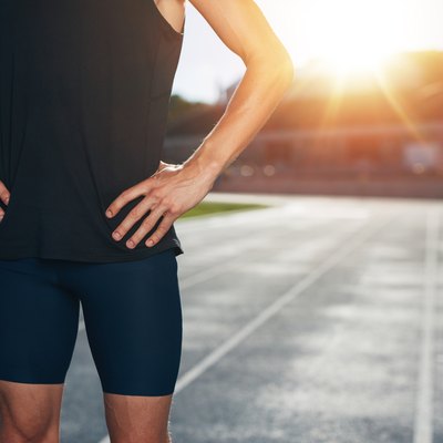 Mid section shot of male athlete standing on race track with his hands on hips on a bright sunlight. Cropped shot of young man runner on athletics running track in stadium.