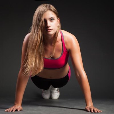 Young girl in sport outfit making push-up on hands and looking at camera on plain black background.