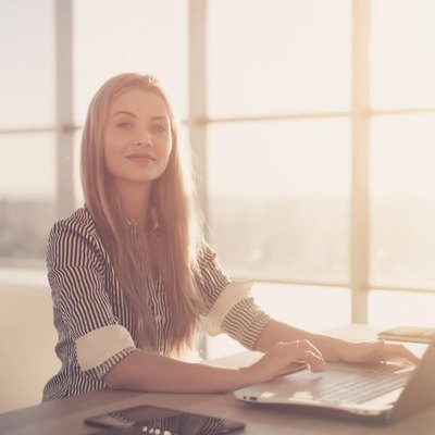 Woman blogging in spacious office using computer on her workplace. Female employee sitting, smiling, looking at camera